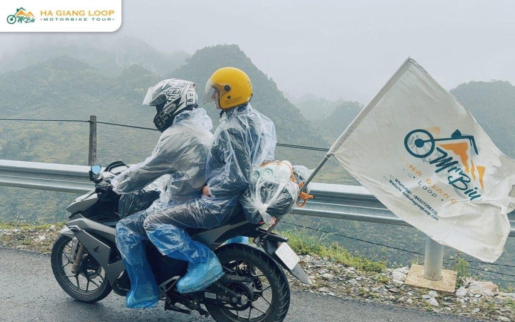 Pillion passenger in a rain poncho holding a Mr. Biu flag